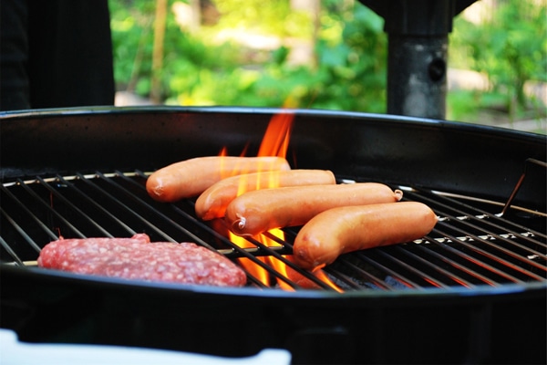 Sausages and a hamburger patty sizzle on the grill with flames and lowcountry greenery in the background.