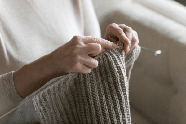 Close-up of hands knitting a ribbed gray fabric with needles, dreaming of a Fripp Island beach vacation.