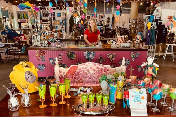 A woman stands behind a colorful gift shop counter filled with art and glassware on a Lowcountry family vacation.
