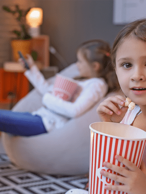 Two young girls on a family vacation eating popcorn and watching a movie at home, one close up, the other in the background.