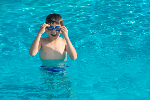 A boy in a swimming pool adjusts his blue goggles while standing in the water.
