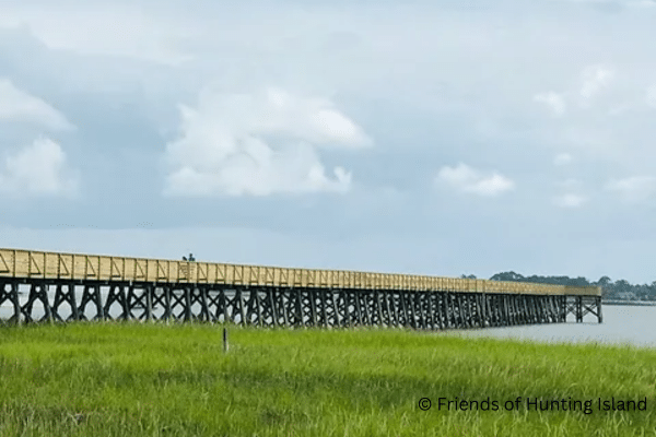 A long wooden pier stretches over the water with green marsh grass in the foreground under a cloudy sky.