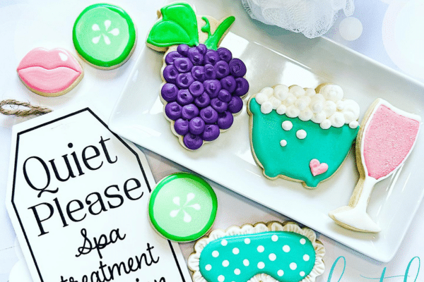 Decorated spa-themed cookies, including grapes, a face mask, wine glass, lips, and bubbles, on a white tray.