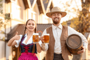 Two people in traditional clothing hold beer mugs and smile outdoors; one gives a thumbs up, the other holds a barrel.