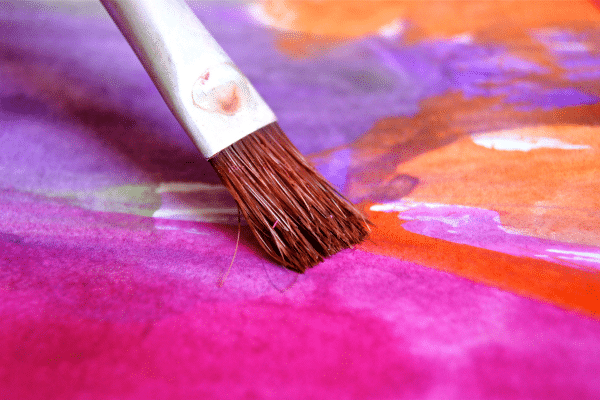 A paintbrush applies bright orange and purple watercolor paint on paper.
