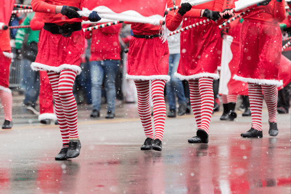 People in Santa costumes and red-and-white striped tights marching in a holiday parade on a snowy street.