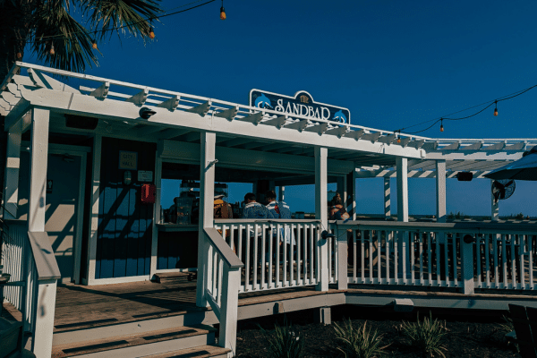 Outdoor view of a seaside restaurant named Sandbar with people sitting inside under a white pergola.