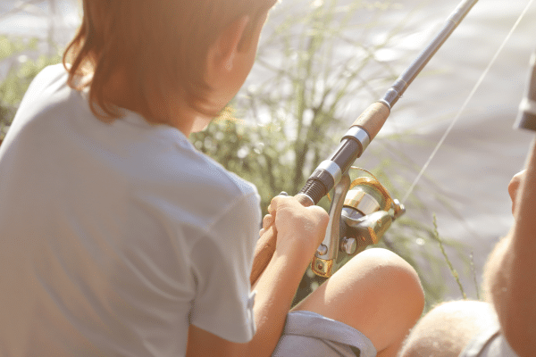 Child holding a fishing rod, sitting by the water with sunlight shining, seen from behind.
