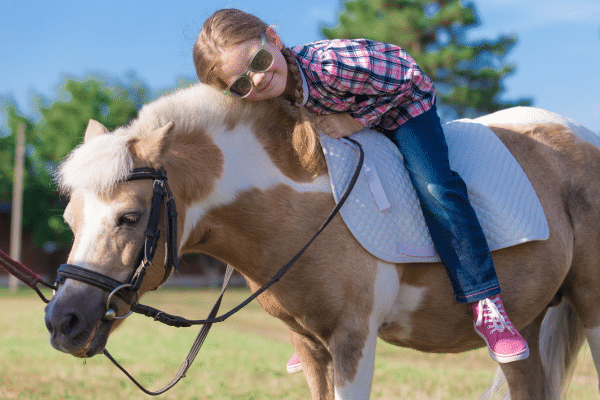 A smiling girl in sunglasses lies on a pony outdoors, wearing a plaid shirt and pink shoes.