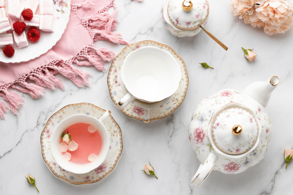 Floral teacups and teapot on marble table with pink cloth, sweets, and small flowers.