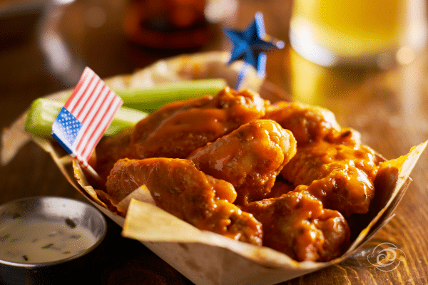 Basket of saucy chicken wings with U.S. flag toothpick, celery sticks, and dipping sauce on a wooden table.