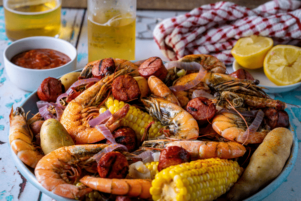 A plate of shrimp, sausage, corn, potatoes, and onions, with drinks, sauce, and lemon halves nearby.