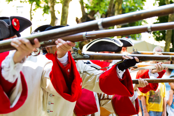 People in historical uniforms hold muskets in a row, aiming forward during a Lowcountry reenactment outdoors.