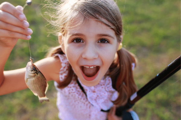 Smiling girl on a beach vacation holding up a small fish she caught, looking excited and surprised.