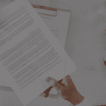 Two people exchange a document over a desk with a notebook, pen, and glass of water nearby.