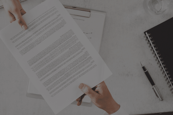 Two people exchange a document over a desk with a notebook, pen, and glass of water nearby.
