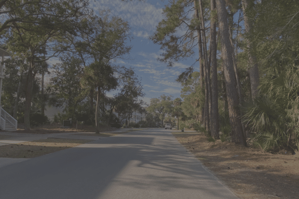 Sunny suburban street lined with tall trees and houses under a blue sky with scattered clouds.