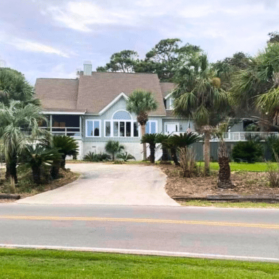 Light blue house with palm trees in front, a driveway, and a lawn, seen from across the street.