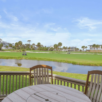 Wooden patio table and chairs overlook a golf course, pond, palm trees, and nearby houses under a blue sky.