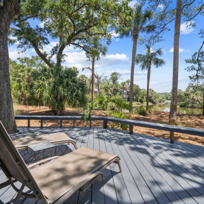 Two lounge chairs on a gray deck overlook palm trees and a pond under a bright blue sky with scattered clouds.