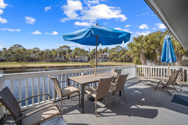 Patio with table, chairs, and blue umbrella overlooks water, ocean point golf course, and houses under a bright blue sky.