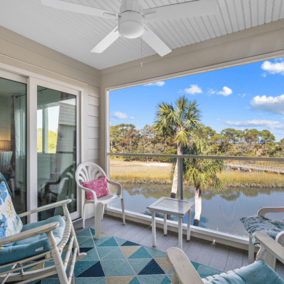Screened porch with chairs, a rug, and ceiling fan overlooking a pond with palm trees and blue sky.