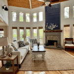 Bright living room with tall windows, a white sofa, fireplace, armchairs, and a jute rug on hardwood floors.