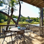 Round table with four chairs on a wooden deck overlooking a pond and trees, with houses in the background.