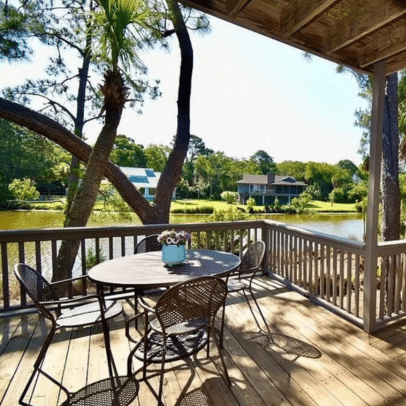 Round table with four chairs on a wooden deck overlooking a pond and trees, with houses in the background.