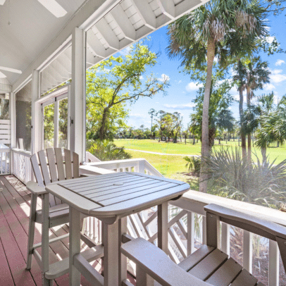 White patio chairs and table on a porch overlook a sunny, green landscape with palm trees.