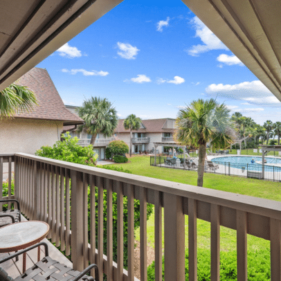 View from balcony overlooking pool, palm trees, and apartment buildings under a blue sky with scattered clouds.
