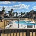 View of a sunny outdoor pool with lounge chairs, palm trees, and buildings in the background.