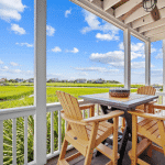 Table and four yellow chairs on a porch overlook green grass and distant beach houses under a blue sky.