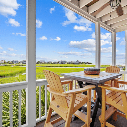 Table and four yellow chairs on a porch overlook green grass and distant beach houses under a blue sky.