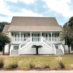 A white house with double front stairs, a porch, and a palm tree in front, set against a cloudy sky.