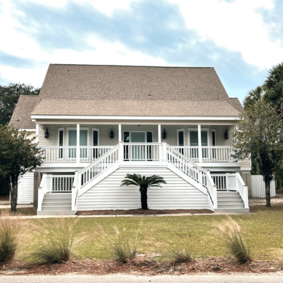 A white house with double front stairs, a porch, and a palm tree in front, set against a cloudy sky.