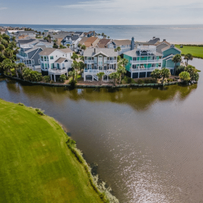Large waterfront houses line a curved shore with a grassy area and ocean in the background.