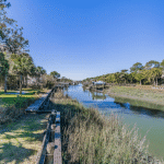 Calm creek lined with wooden docks, tall grass, and trees under a clear blue sky.