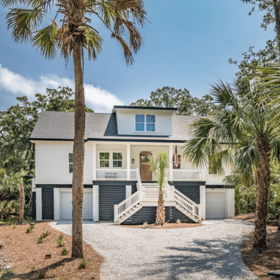 Two-story house with a front porch, palm trees, and gravel driveway under a blue sky.