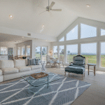Bright living room with large windows, white sofa, glass coffee table, and view of green lawn and water.