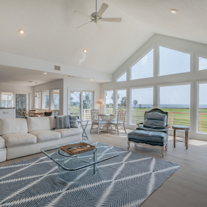 Bright living room with large windows, white sofa, glass coffee table, and view of green lawn and water.