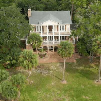 Aerial view of a two-story southern-style house with large porch, surrounded by trees and palm trees.