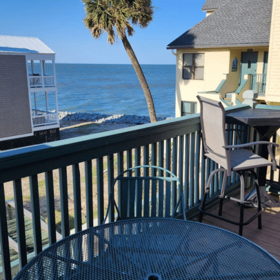 Round table and chairs on a balcony overlook the ocean, with palm tree and buildings nearby.