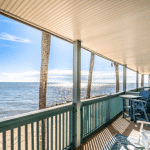 Beachfront porch with blue railing, chairs, and table overlooking the ocean under a sunny sky.