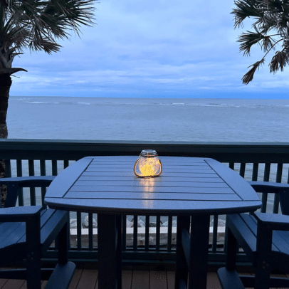 A lit lantern sits on a table by the ocean, with palm trees and a cloudy blue sky in the background.