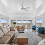 Bright living room with large windows, neutral sofas, blue chairs, and view of outdoors under a white vaulted ceiling.