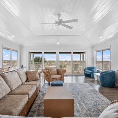 Bright living room with large windows, neutral sofas, blue chairs, and view of outdoors under a white vaulted ceiling.
