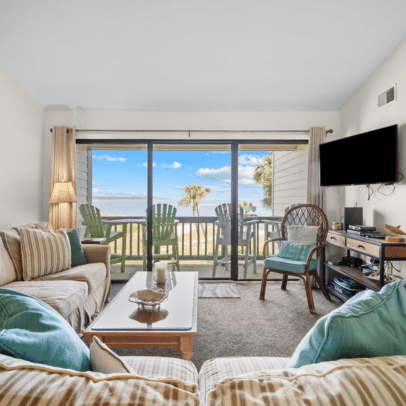 A cozy living room with sofas, a TV, and a balcony overlooking palm trees and the ocean.