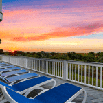 Blue lounge chairs on a deck overlook greenery and a vibrant sunset with pink, orange, and yellow skies.