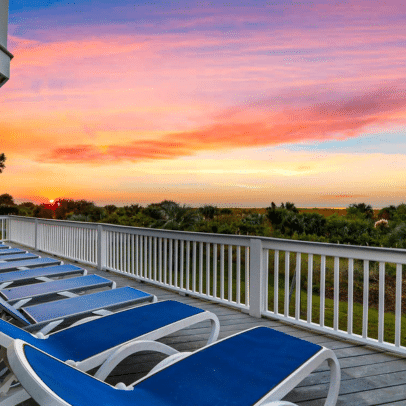 Blue lounge chairs on a deck overlook greenery and a vibrant sunset with pink, orange, and yellow skies.